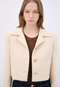 Beige kort jacka i texturerat tyg med slagkrage, två framfickor och två gyllene knappar. Bärs över en brun topp.