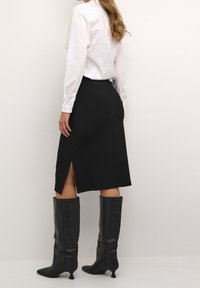 Schwarzer Bleistiftrock mit einem seitlichen Schlitz, kombiniert mit kniehohen schwarzen Lederstiefeln. Das Oberteil ist ein tailliertes weißes Hemd mit Knöpfen.