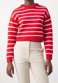 Pull en tricot rayé rouge et blanc avec un col rond et un design cropped. Un pantalon couleur crème est visible. Tissu texturé.