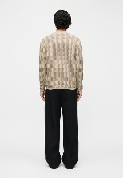 Personne aux cheveux courts et bouclés vue de dos, portant un haut beige texturé à manches longues et un pantalon large noir et ample.