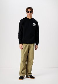 Zwart sweatshirt met een logo, olijfgroen wijde broek en zwarte sneakers. Model draagt een zonnebril en staat tegen een witte achtergrond.