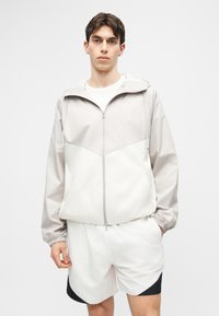 Νέος άνδρας που φορά ελαφρύ γκρι φούτερ με φερμουάρ και κουκούλα και λευκά αθλητικά σορτς με μαύρες λεπτομέρειες, στέκεται μπροστά σε μονόχρωμο φόντο.