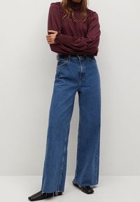 Blauwe denim wide-leg jeans met een versleten zoom, gecombineerd met een kastanjebruine top met lange mouwen die textuurelementen en gerimpelde details heeft.