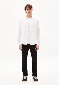 Chemise blanche à manches longues, portée sur un T-shirt blanc. Jean noir et baskets noires avec des accents crème. Design simple, sans motifs.