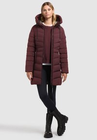 Femeie îmbrăcată cu o jachetă lungă din material quilted burgundy peste un hanorac, leggings negri și cizme groase negre, stând în fața unui fundal deschis.