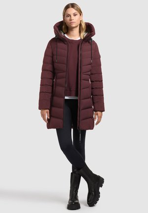 Nő hosszú bordó steppelt kabátot visel, alatta pulóver, fekete leggings és fekete chunky csizma, világos háttér előtt áll.