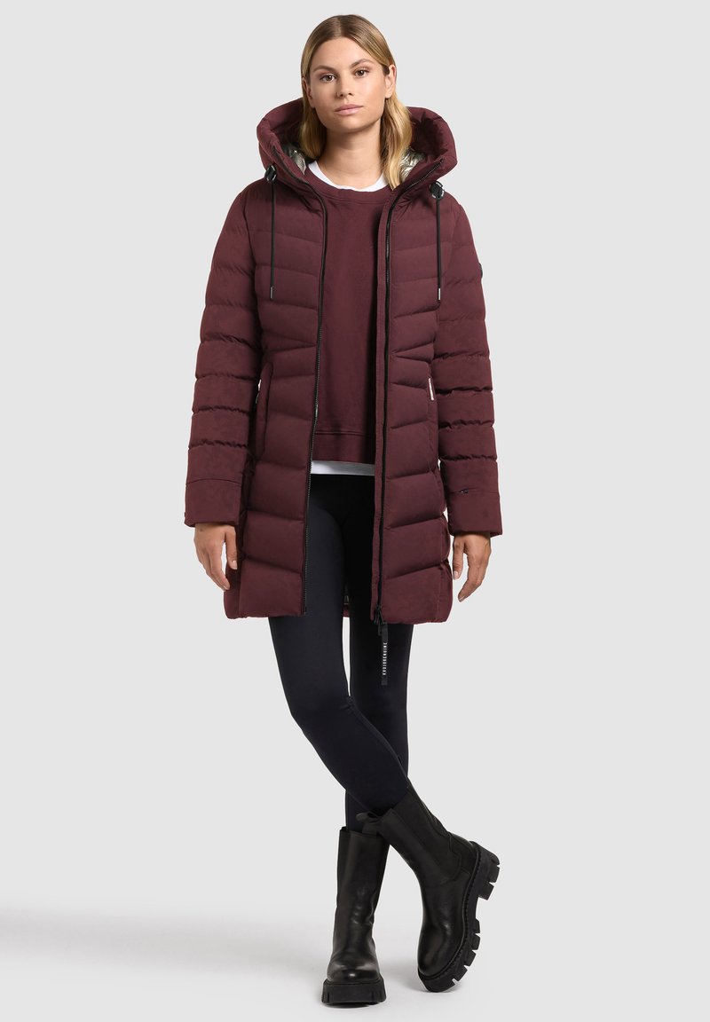 Femeie îmbrăcată cu o jachetă lungă din material quilted burgundy peste un hanorac, leggings negri și cizme groase negre, stând în fața unui fundal deschis.