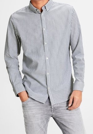 Mężczyzna ubrany w szarą i białą pionowo prążkowaną koszulę na guziki z długimi rękawami oraz jasnoszare dżinsy, z rękami w kieszeniach.