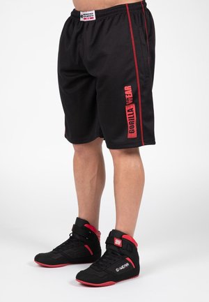 Shorts de sport noirs avec des rayures latérales rouges et le logo "GORILLA WEAR". Portés avec des chaussures de sport montantes noires avec des accents rouges.