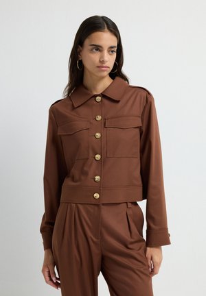 Vrouw met lang zwart haar, gekleed in een bruine knopenjasje en bijpassende geplooide broek, staand tegen een effen lichte achtergrond.