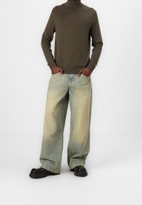 Zielona oliwkowa sweter z golfem, sparowany z jasnoniebieskimi spodniami o szerokich nogawkach. Czarne buty na grubej podeszwie. Minimalistyczny design, gładka tekstura.