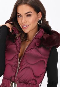 Burgundowa kamizelka puchowa z futrzanym kapturem, zamek błyskawiczny, pikowany materiał oraz pasek w talii z okrągłym metalowym akcentem.