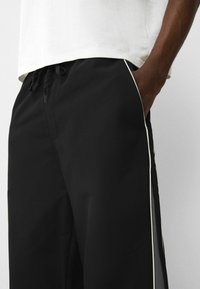 Persoon in zwarte broek met witte piping aan de zijkant en een wit shirt, hand in de rechterzak gestoken.