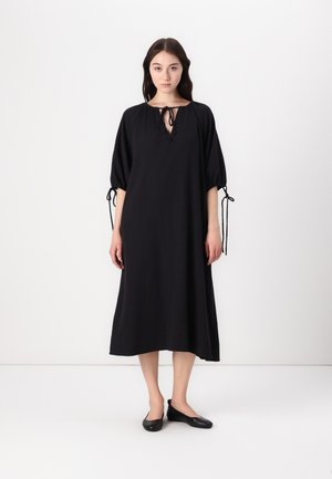 Black midi dress with balloon sleeves, drawstring neckline, and flowing silhouette. Smooth fabric with a soft texture. Worn with black flats.