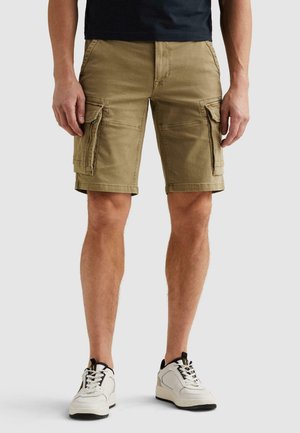 Man draagt beige cargoshorts met zijzakken en witte sneakers, staand tegen een effen lichte achtergrond.