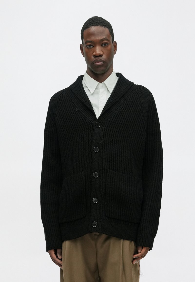 Cardigan a coste nera con colletto a scialle, dotato di cinque bottoni e due tasche frontali. Indossato sopra una camicia bianca con colletto.