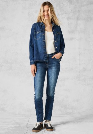 Denim jacket with two chest pockets, buttoned front, and long sleeves, paired with dark blue jeans and black shoes with white laces.