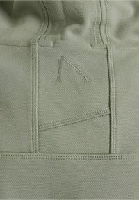 Gros plan d'un sweat à capuche vert en tissu avec des coutures visibles et un logo triangulaire discrètement cousu au dos, près de la capuche.
