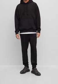 Hoodie nera con logo in rilievo, ampia vestibilità, tasca frontale, abbinato a pantaloni della tuta neri. Le sneakers presentano suole testurizzate e lacci.