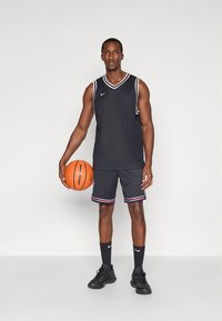 Czarny koszulkowy tank top do koszykówki z białymi akcentami, zestawiony z czarnymi szortami z czerwono-białymi paskami. Trzyma pomarańczową piłkę do koszykówki.