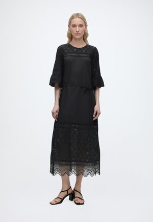 Naine kannab musta midi pikkusega kleiti, millel on silmusega pitsdetailid, kolmveerandvarrukad volangidega, vöökohalt seotav ja mustad kontsakingad, seisab valgel taustal.