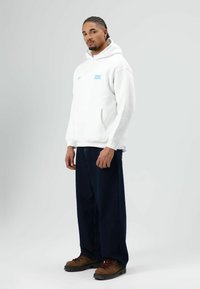 Witte hoodie met een blauw logoplaatje, gecombineerd met donkere wijde jeans en bruine veterlaarzen. Eenvoudig ontwerp, zachte textuur en een losse pasvorm.