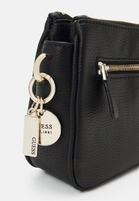 Sort læderhåndtaske med tekstureret overflade, guldfarvet hardware, lynlås og et vedhæng med mærkenavnet "GUESS".