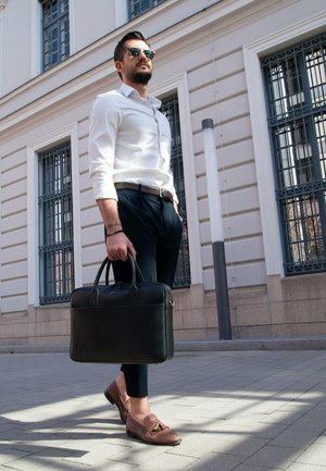 Black leather briefcase held by a man in a white shirt and dark pants, with brown loafers featuring tassel details. Modern urban setting.