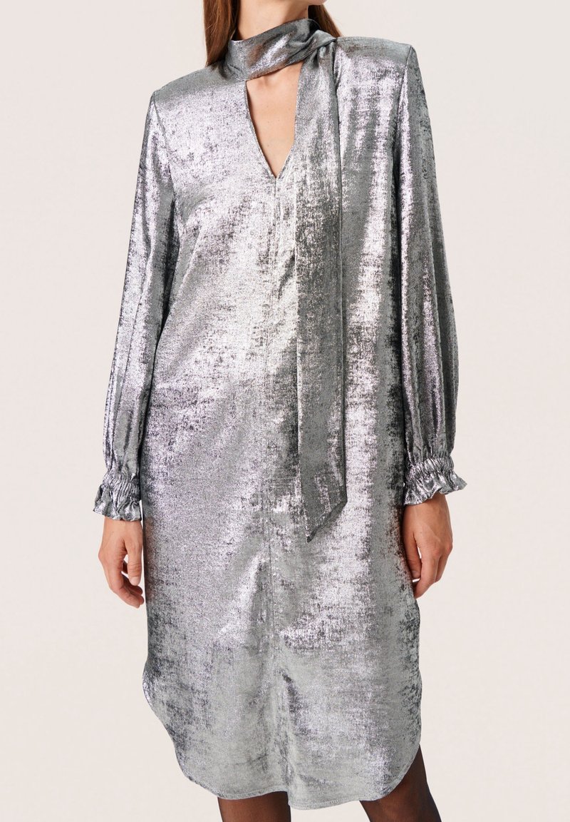 Vestido metálico plateado con mangas largas, escote en V y detalle de cuello atado. Presenta puños fruncidos y un corte holgado. Textura suave.