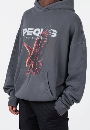Ciemnoszara bluza z kapturem z dużą grafiką orła i napisem "PEQUS" u góry, noszona przez osobę z rękami częściowo w kieszeniach.