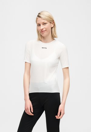 Šviesiaplaukė moteris, vilkinti aptemptą baltą trumpomis rankovėmis megztinį su užrašu „sportful“ ir juodus kelnes, stovinti priešais vienspalvį foną.