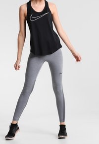 Top senza maniche Nike nero con logo bianco, abbinato a leggings grigi con una striscia laterale e logo Nike nero, indossato con sneaker nere.
