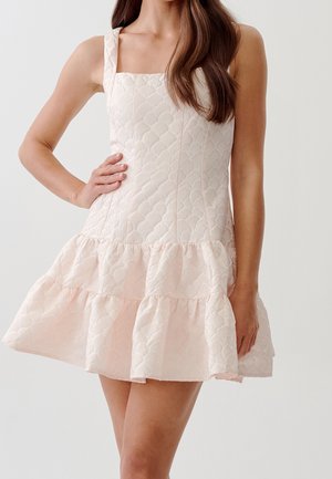 Nainen pukeutuneena vaaleanpunaiseen, hihattomaan minimekkoon, jossa on teksturoitu merkkikuvio ja röyhelöidyt helmat, poseeraa yhdellä kädellä lantiolla.