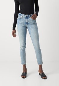 Jeans in denim azzurro chiaro con un design aderente e stile a cinque tasche. Presentano una vita alta e un taglio alla caviglia. Sandali neri a scivolo completano il look.