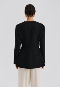 Fekete texturált blézer strukturált sziluettel, karcsúsított derékkal és hosszú ujjakkal. Feltűnő gombdíszítés a mandzsettákon. Tejszínszínű nadrág.