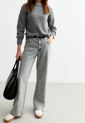 Vrouw met een grijze trui, lichtgrijze wijde jeans, witte sneakers, houdt een zwarte handtas vast, staande tegen een effen witte achtergrond.