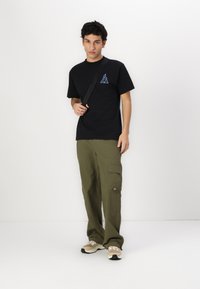 T-shirt en coton noir avec logo triangulaire bleu, associé à un pantalon cargo vert olive avec poches latérales et une finition texturée.
