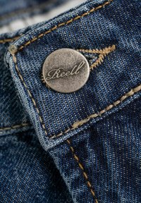 Tejido de mezclilla azul con un botón de metal visible grabado "Reell." Presenta costuras amarillas y una superficie texturizada con diferentes tonalidades.