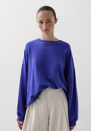 Pulover oversized mov, cu guler rotund și umeri lăsați, asortat cu pantaloni largi bej deschis, având un material texturat.