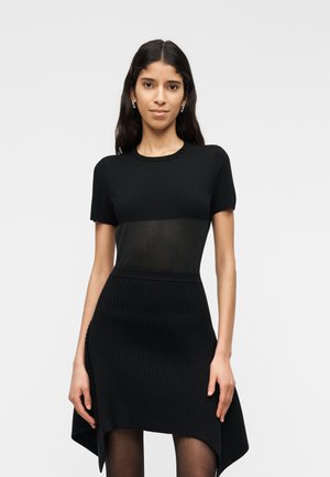 Kvinde iført en tætsiddende sort kjole med korte ærmer og en ribbet, asymmetrisk kant, stående mod en ensfarvet lys baggrund.