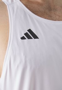 Hvid træningstanktop med en glat tekstur, rund halsudskæring og et fremtrædende sort Adidas-logo øverst til venstre.