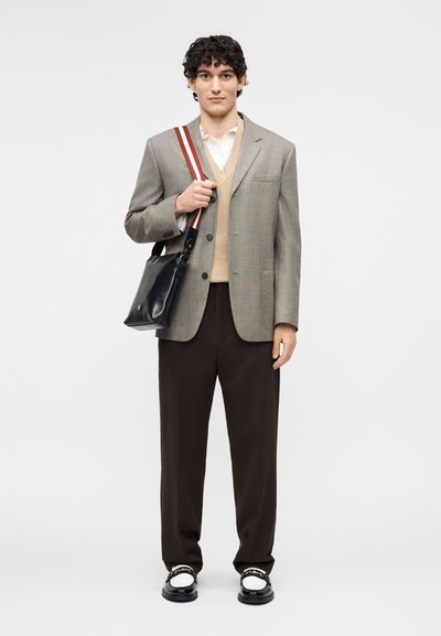 Young man standing wearing gray blazer, beige sweater vest, white shirt, dark pants, black-and-white loafers, and carrying black shoulder bag.