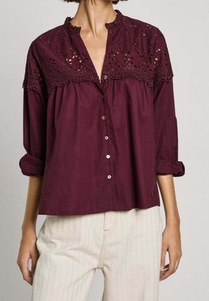 Kvinde iført en bourgognefarvet knap-op bluse med blomsterblonde på yoke og ærmer rullet op, kombineret med lyse beige bukser.