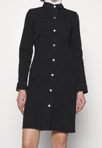 Černé denimové šaty s dlouhými rukávy, zapínáním na knoflíky, límcem a dvěma náprsními kapsami. Obsahují bílé knoflíky a mají přiléhavý siluetu.