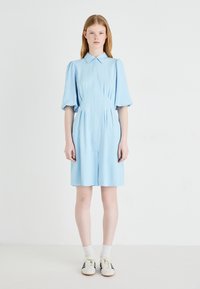 Robe bleu clair avec un devant à boutons, des manches courtes bouffantes et des détails plissés. Associée à des chaussettes blanches et des baskets à rayures noires.