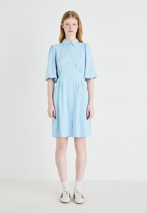 Robe bleu clair avec un devant à boutons, des manches courtes bouffantes et des détails plissés. Associée à des chaussettes blanches et des baskets à rayures noires.