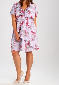 Kukallinen mekko vaaleanpurppurassa, jossa on röyhelödetaljeja, lyhyet hihat ja kapea vyötärö. Yhdistetty nudenvärisiin korkokenkiin.