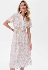 Witte jurk met gouden bloemenpatroon, korte mouwen, V-hals en elastische taille. De gegolfde zoom voegt detail toe aan het halflange ontwerp.