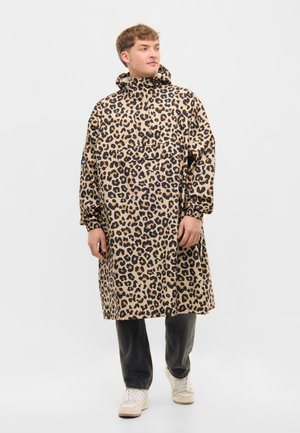 Férfi, aki hosszú, zsebes, kapucnis, leopárdmintás kabátot, fekete nadrágot és fehér sportcipőt visel, egyszerű háttér előtt áll.