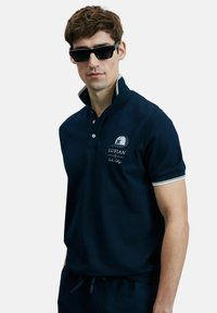 Marineblauwe polo met witte accentstrepen op de kraag en mouwen; heeft een logo patch en geborduurde merknaam.